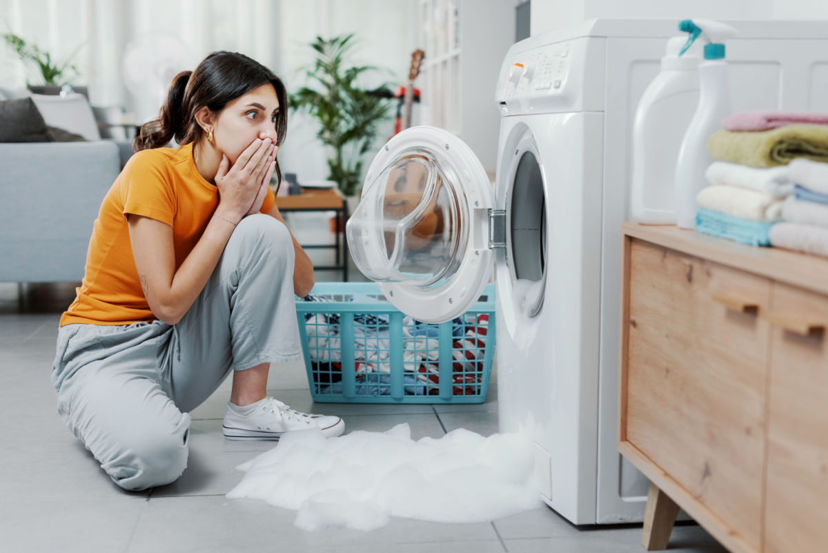 A shocked woman looking at a washing machine that's overflowing with suds.