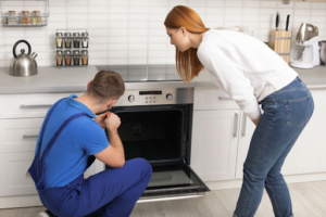 Repairman working on broken oven as female homeowner watches