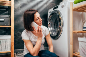 A woman sitting next to a clothes dryer using a phone to call for repair