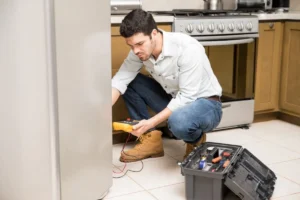 Male technician in white shirt and jeans kneeling behind a refrigerator as he tests a broken component