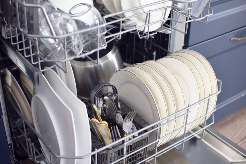 an open dishwasher full of dishes (square plates, round plates, silverware, wine glasses, and small dessert plates)