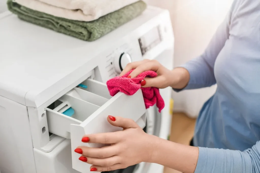 a woman's hand holding a pink rag as she wipes down the detergent dispenser on the washing machine