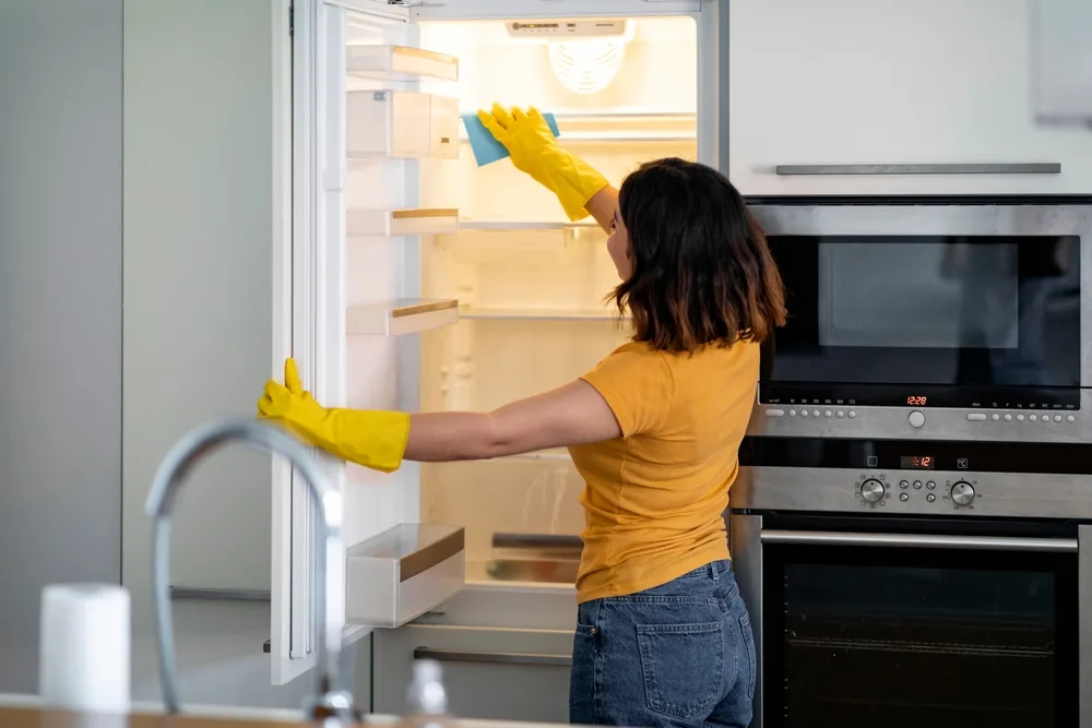 a woman with yellow gloves wiping down the interior of an empty refrigerator
