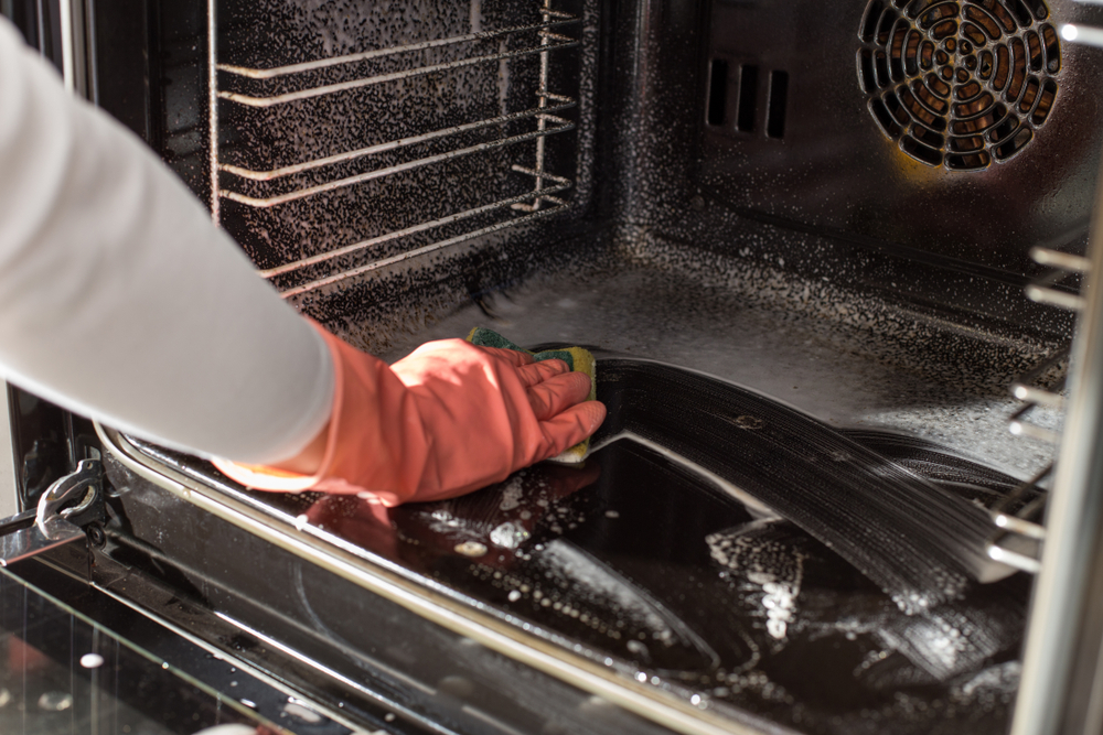 a woman's hand in a pink glove and holding a green and yellow sponge scrubbing at the interior of the oven, where oven cleaner is sprayed
